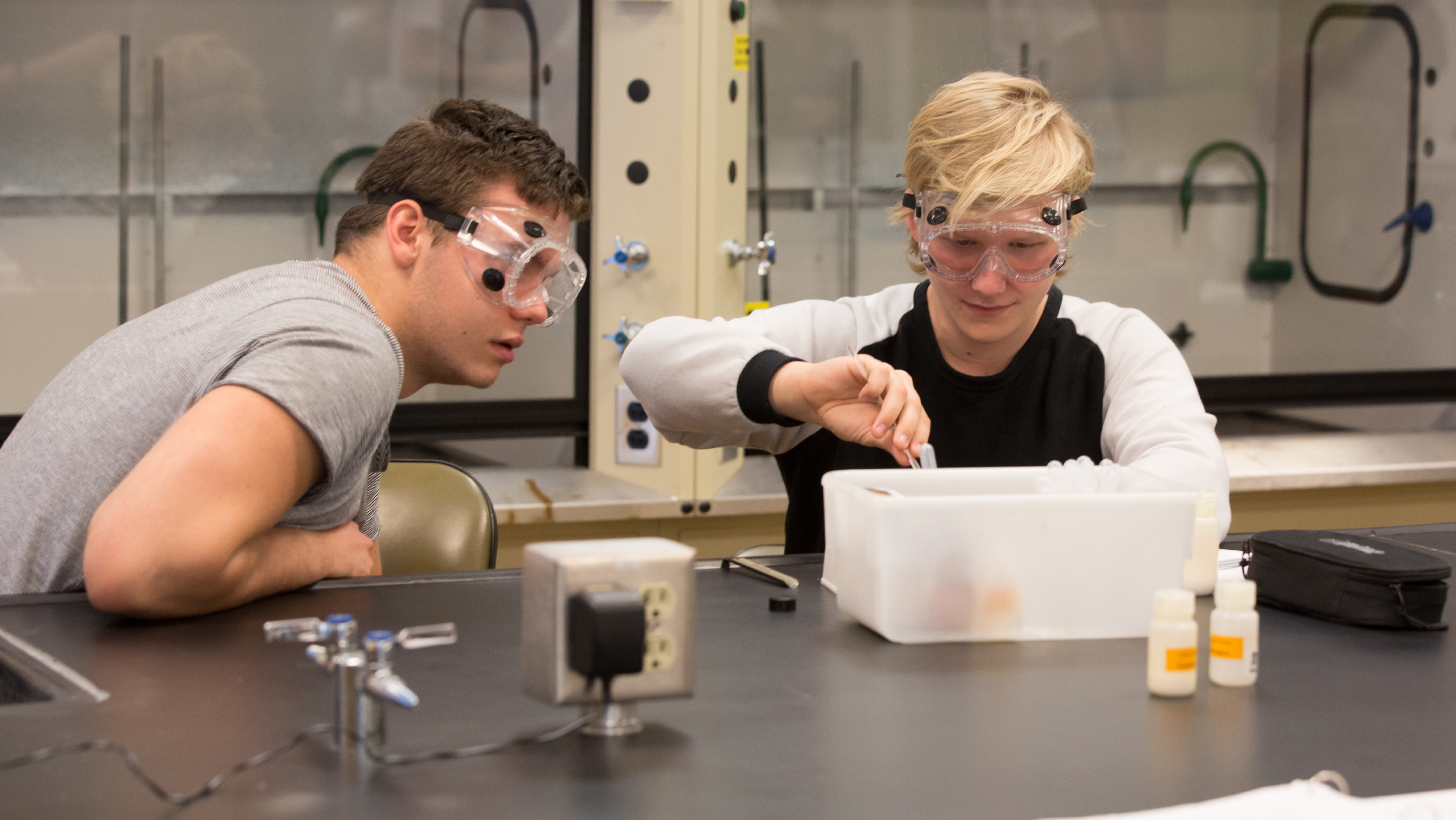 Two students wearing lab safety goggles conducting an experiment at a chemistry lab bench.