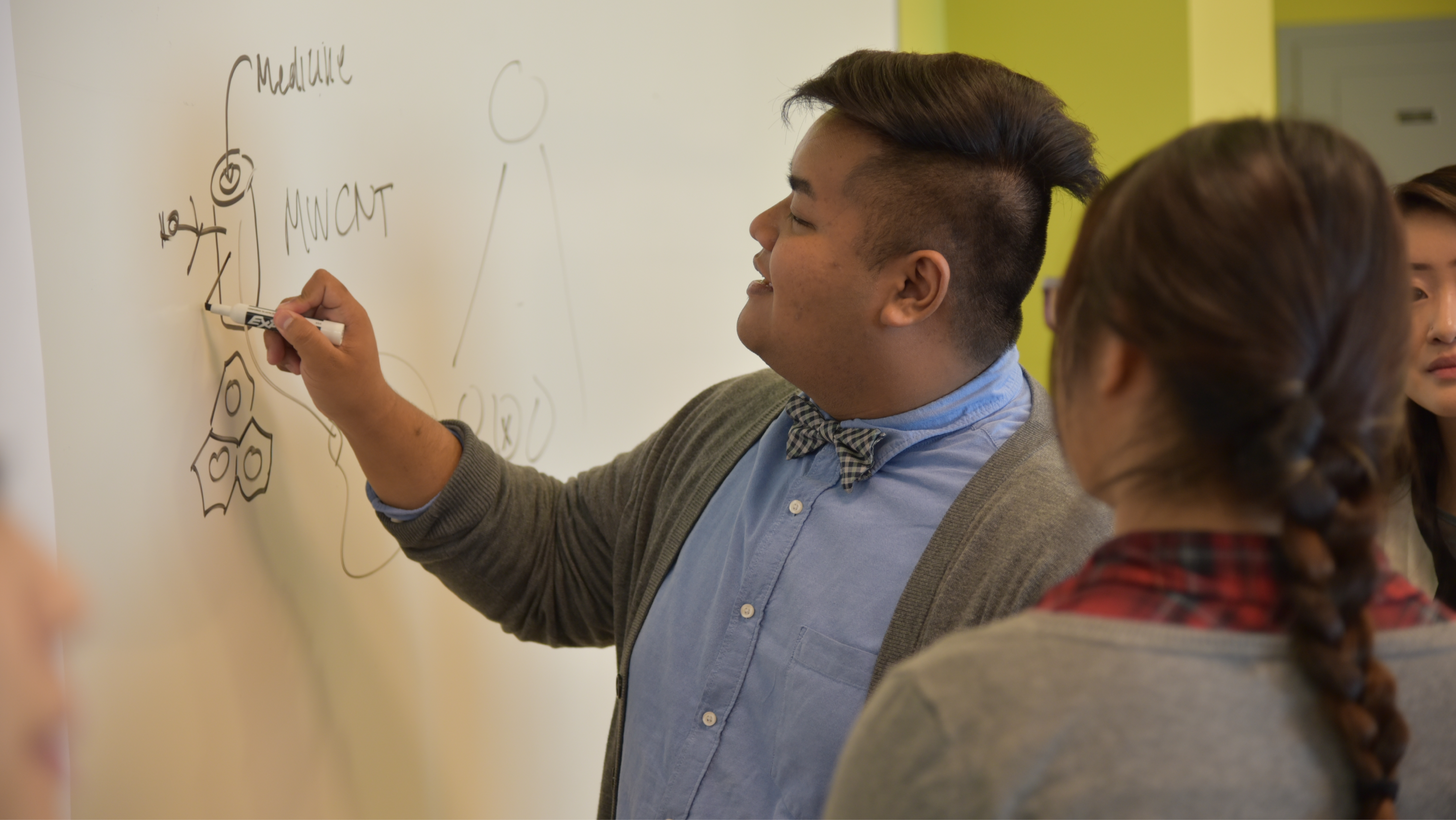 A student in a bowtie drawing molecular structures and diagrams on a whiteboard for classmates.