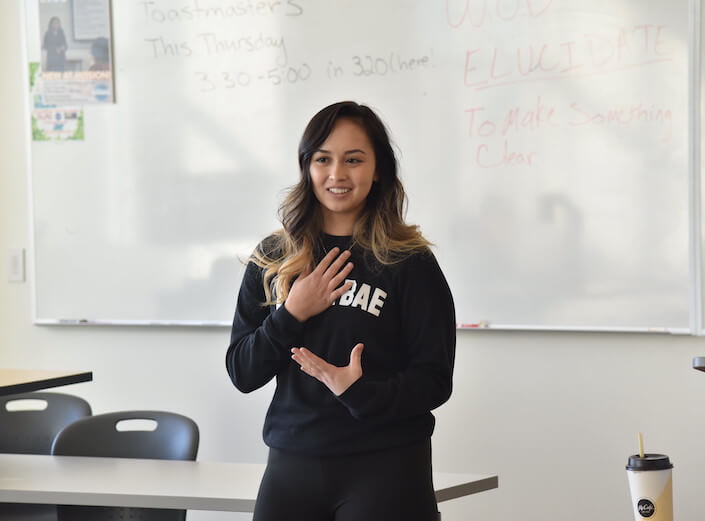 Young woman in a dark blue sweatshirt that reads "Boba Bae" delivers a speech in her classroom. She has wavy dark hair with blonde highlights and is of Asian descent. She gestures with her hands.