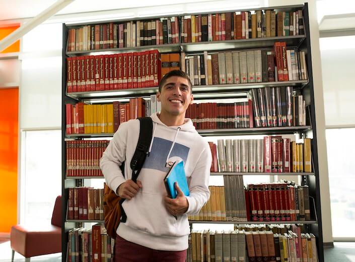 Latinx boy in white hoodie holds a book and poses in the library stacks.