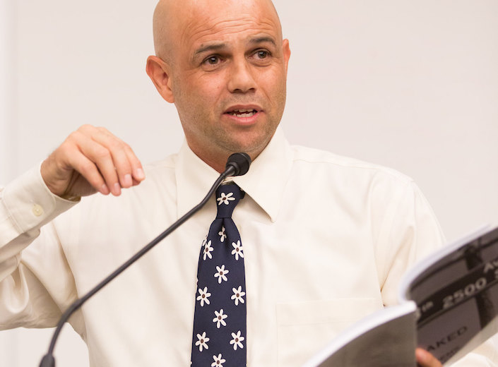 Donnelle McGee reads from one of his published books. He has a shaved head and wears a white shirt and tie.