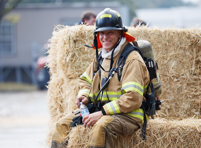 Fire fighter smiles in uniform. He sits on a bale of hay and is a white man.