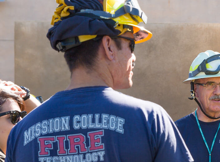 The back of a fire fighter's head is pictured, he wears a yellow helmet and a blue t-shirt that reads "Mission College Fire Technology"