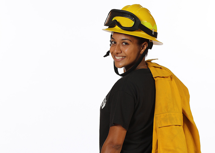 Fire Fighter Academy. A young Latinx fire fighter turns back toward the camera. She has a yellow uniform jacket draped over her shoulder and a yellow fire fighter helmet with goggles on it. 
