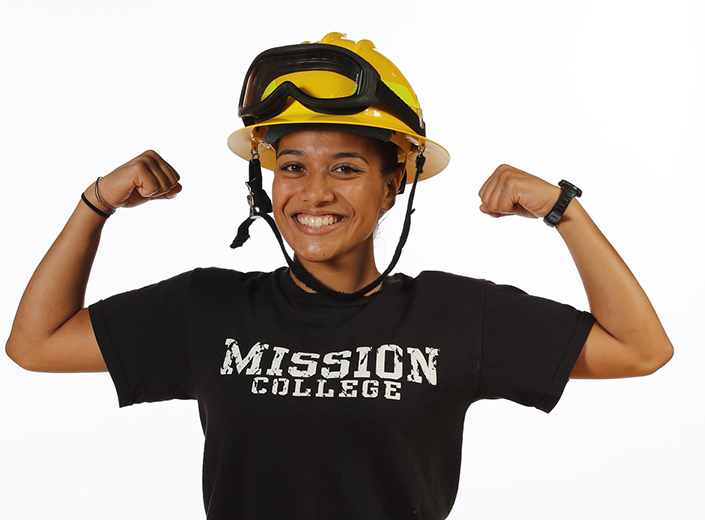 Young Latinx Fire Tech student poses in a black Mission Colelge Tshirt and yellow fire helmet. She has her arms lifted up in a "strong man pose"