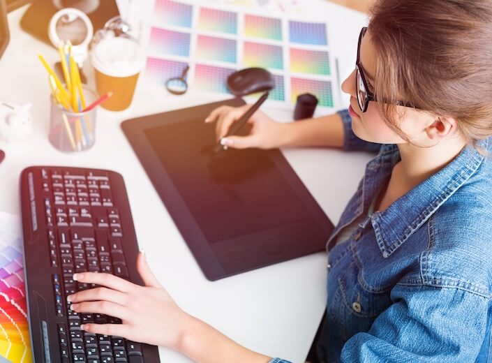 Graphic Design student works at a desk. She is surrounded by colorful papers and wears black plastic glasses.