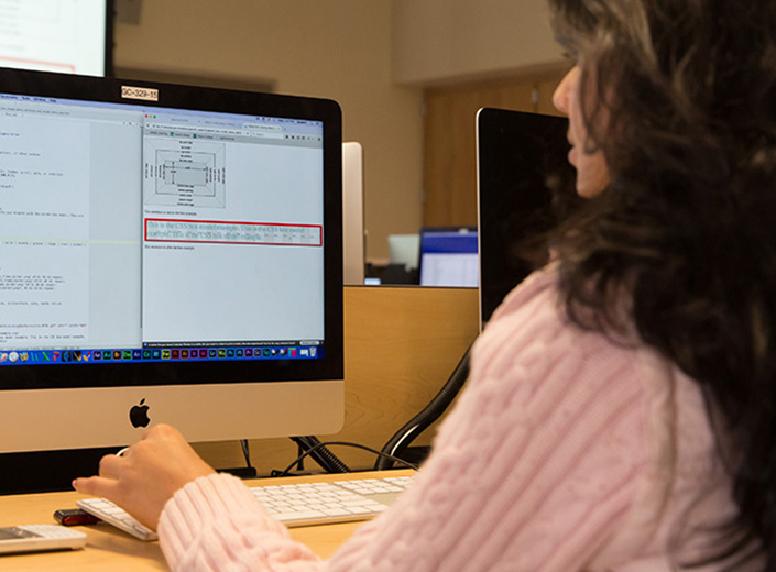 Young woman with dark wavy hair and pink sweater works at a desktop computer using CSS.