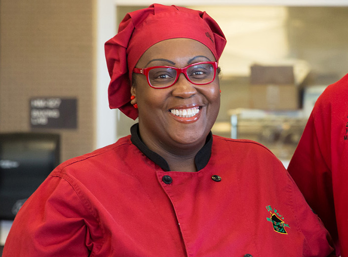 Adult student in Hospitality program wears red chef's uniform. She is Black and wears thick-rimmed plastic glasses. 