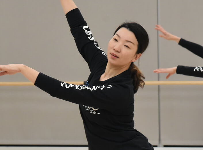 Ballet dancer. A young woman of Asian descene wears a long-sleeved black shirt and raises her arms to the left in a dance move. She dances in a studio with mirrors.