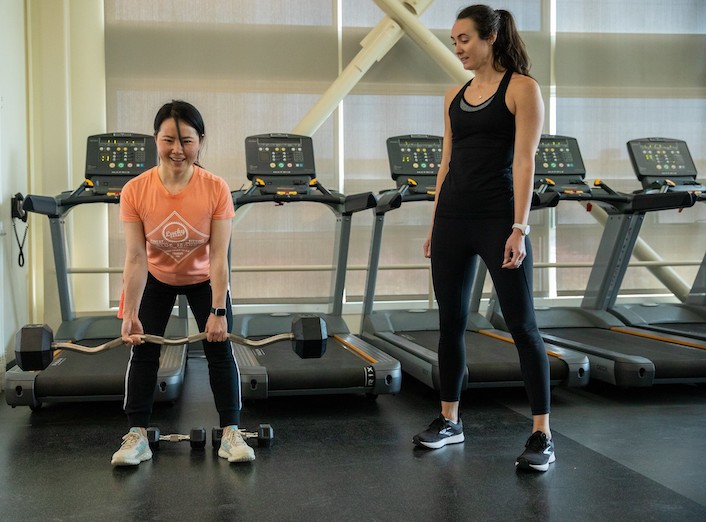 Kaitlin Ferguson, a toned/fit white woman with dark hair in form-fitting workout clothes instructs a female student of Asian descent on how to do a squat lift with a barbell.