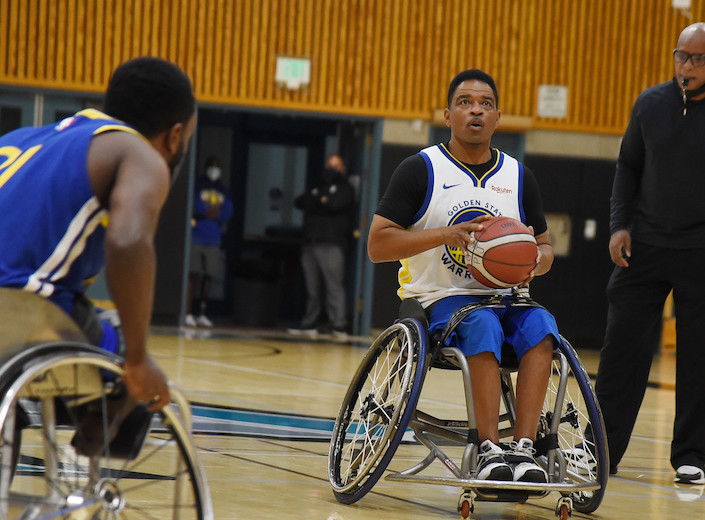 A man in a wheelchair aims at the basketball hoop as he prepares to score a goal. A referee stands besides him with a whistle in his mouth.