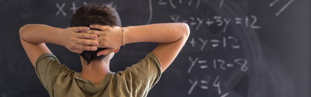 A male student with short brown hair crosses his arms behind his head and contemplates a math problem at the blackboard in his classroom.