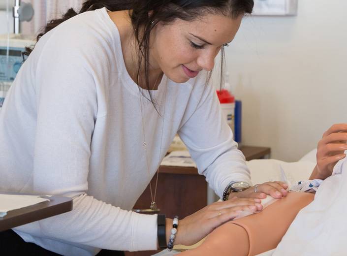 Nursing instructor demonstrates a technique on a dummy in a simulation lab. She has long dark hair and wears a long-sleeved shirt.