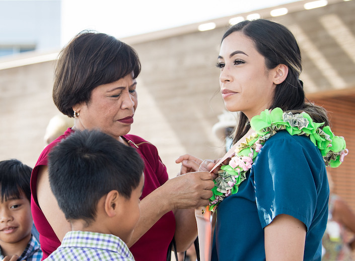 RN pinning ceremony. Student with long black hair wears her blue scrubs and her mother puts a flower leigh around her neck.