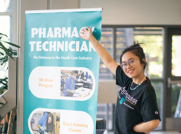 Young woman of Asian descent with a black bun and navy blue "Mission College" t-Shirt gestures toward a tall standing poster advertising the Pharm Tech program.