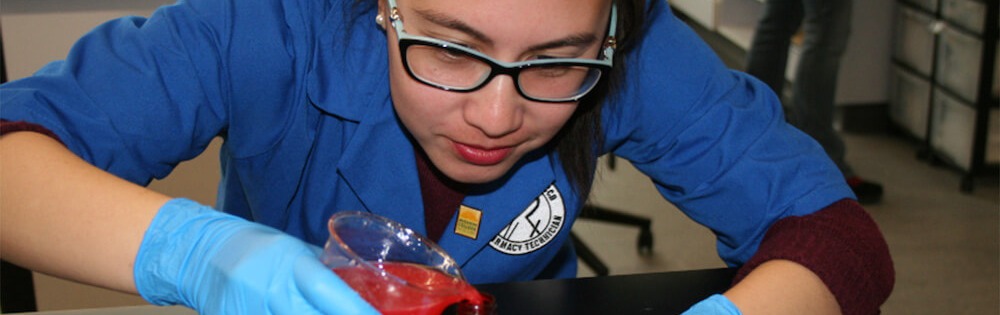 Pharmacy Technican student in blue scrubs and glasses leans over a lab counter to do work with a beaker. She wears gloves, has dark hair pulled back off her face and glasses with dark frames.