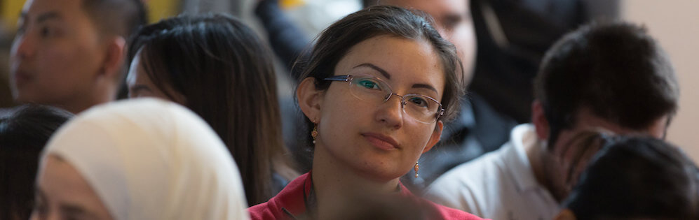 Young woman with dark hair and glasses is seated in the audience at a college event. She looks forward toward the stage (out of sight.)