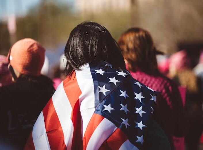 Woman with dark hair marches draped in the American flag. She is seen from behind.