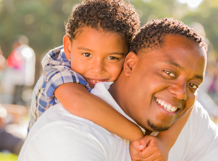 African-American dad and son. The young son throws his arms around dad's neck. They are outside in a sunny park.