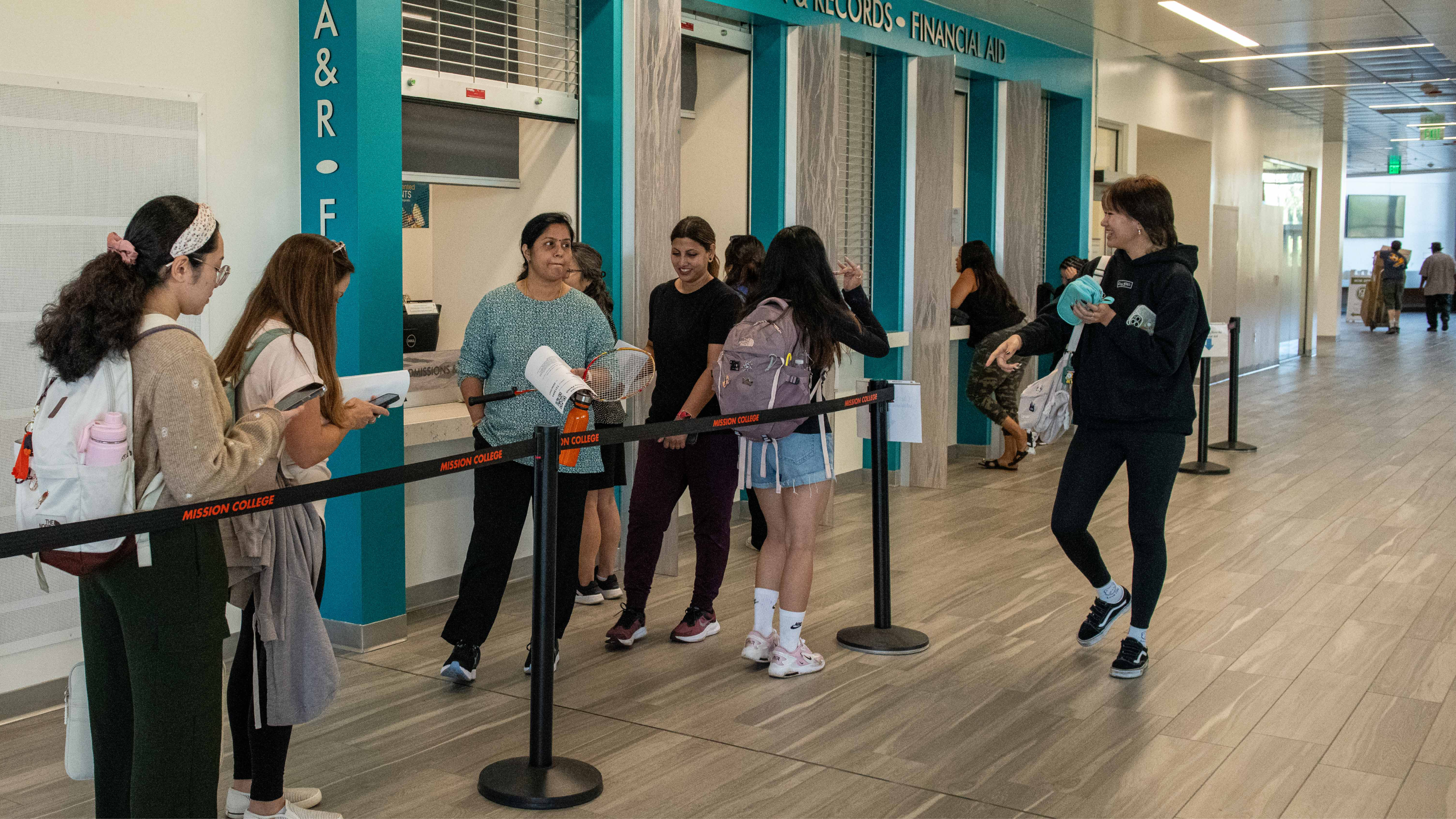 Students waiting in line behind a stanchion near the Admissions and Records counter inside a modern campus building.