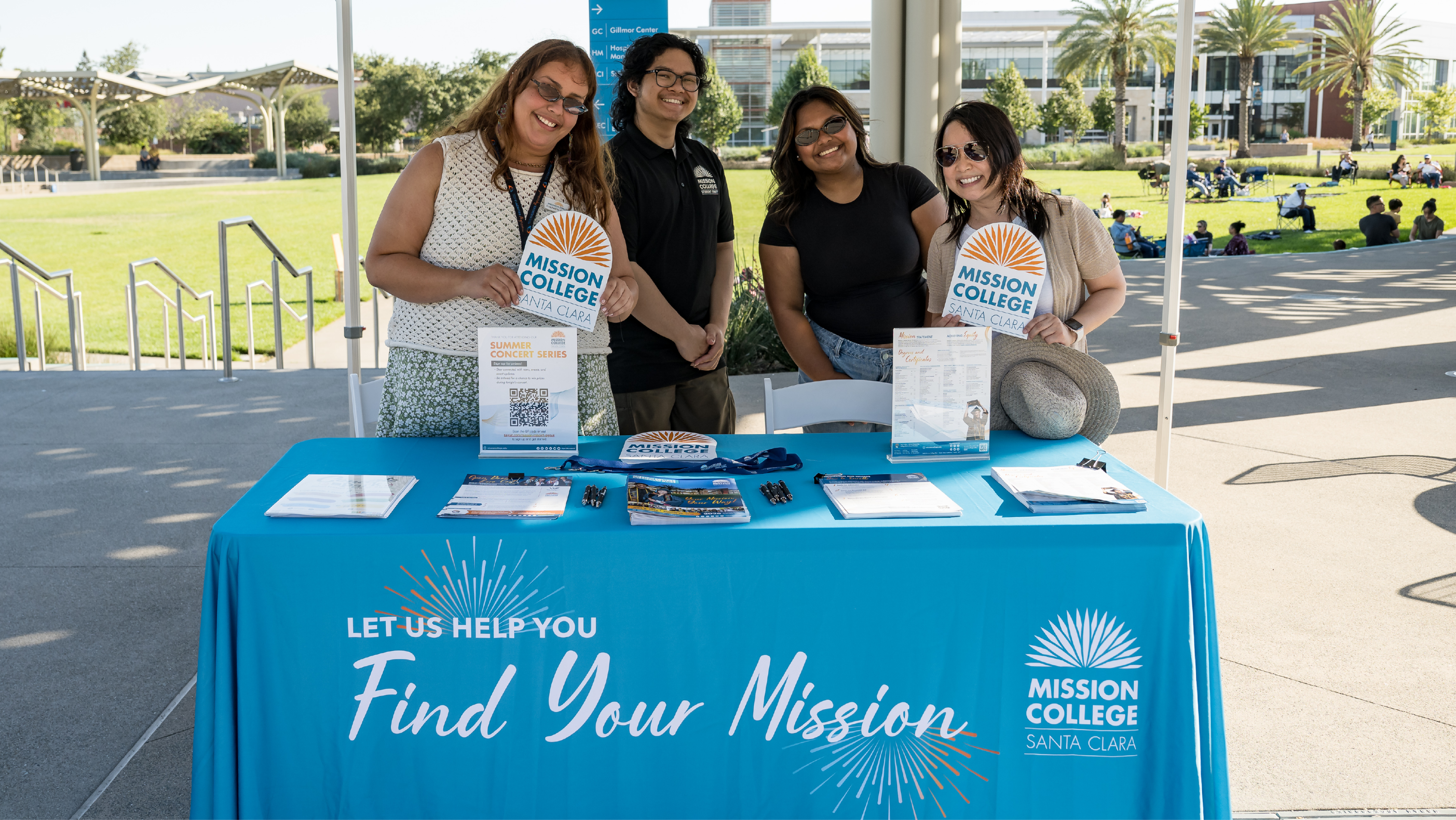 Four smiling Mission College staff members standing behind a blue "Find Your Mission" branded table at an outdoor event.