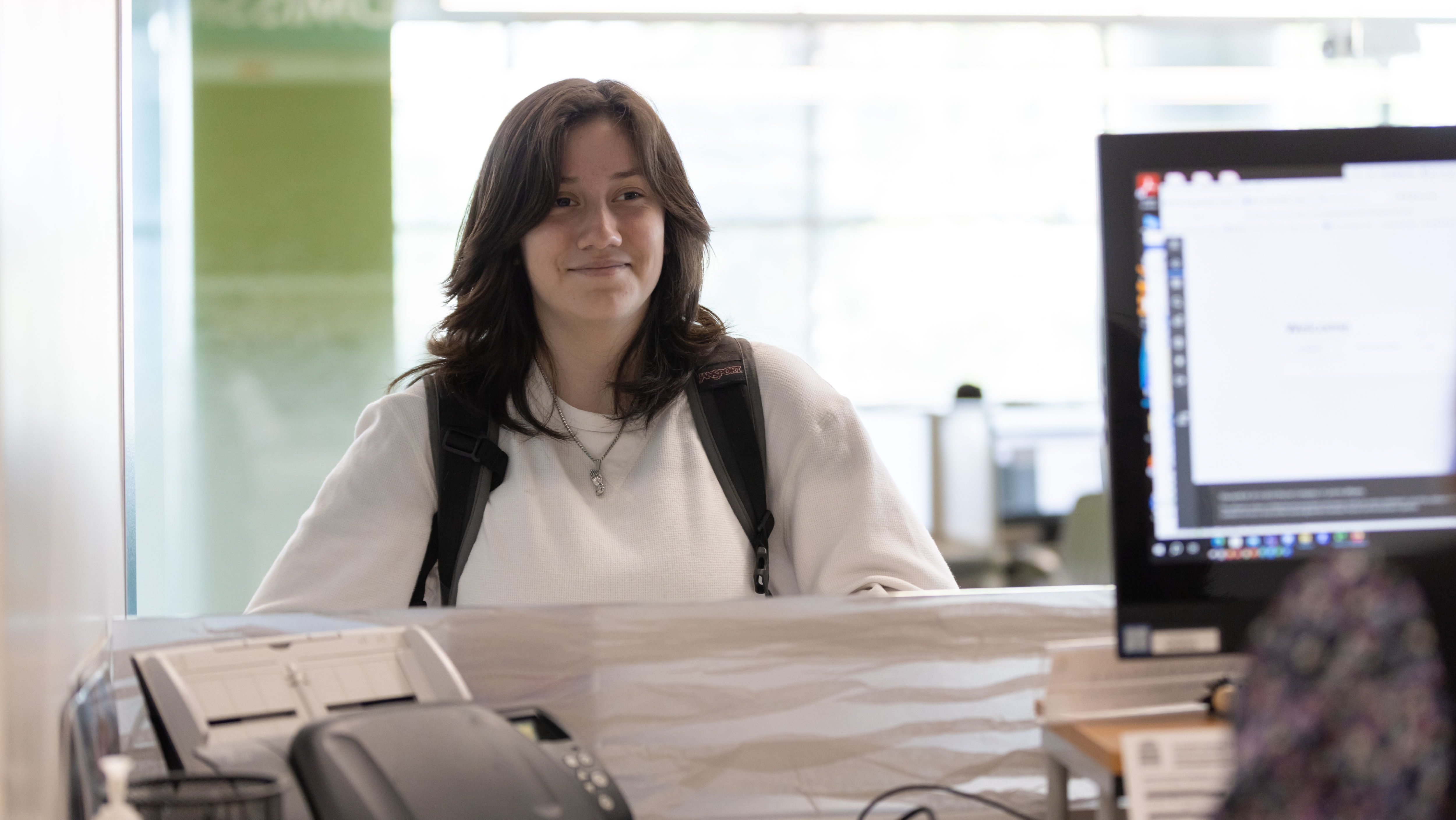 A smiling student wearing a backpack standing at a front desk counter across from a computer screen.
