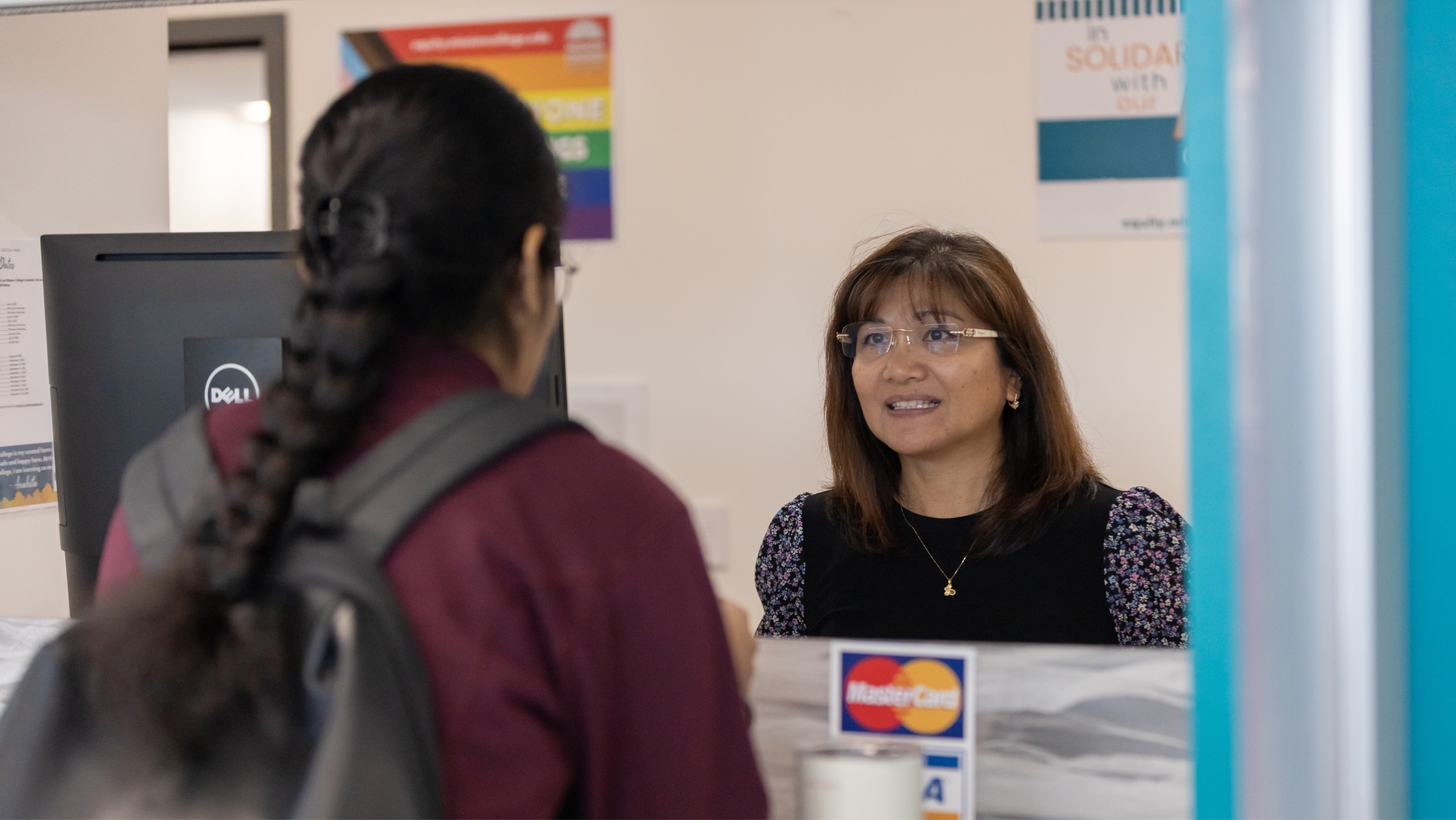A staff member wearing glasses smiling and speaking with a student carrying a backpack at an intake counter.
