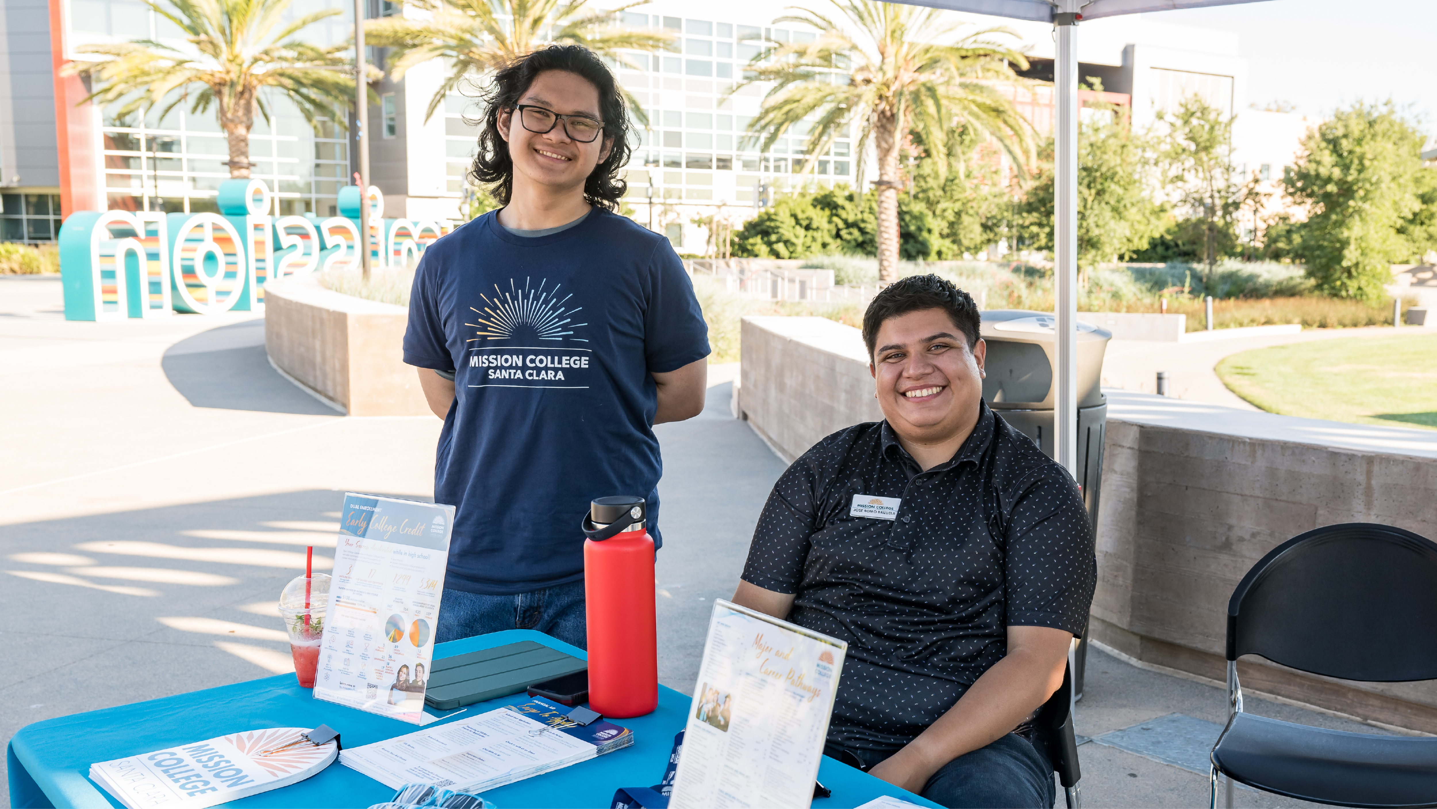 Two student representatives smiling behind a blue Mission College information table set up outdoors in a plaza.