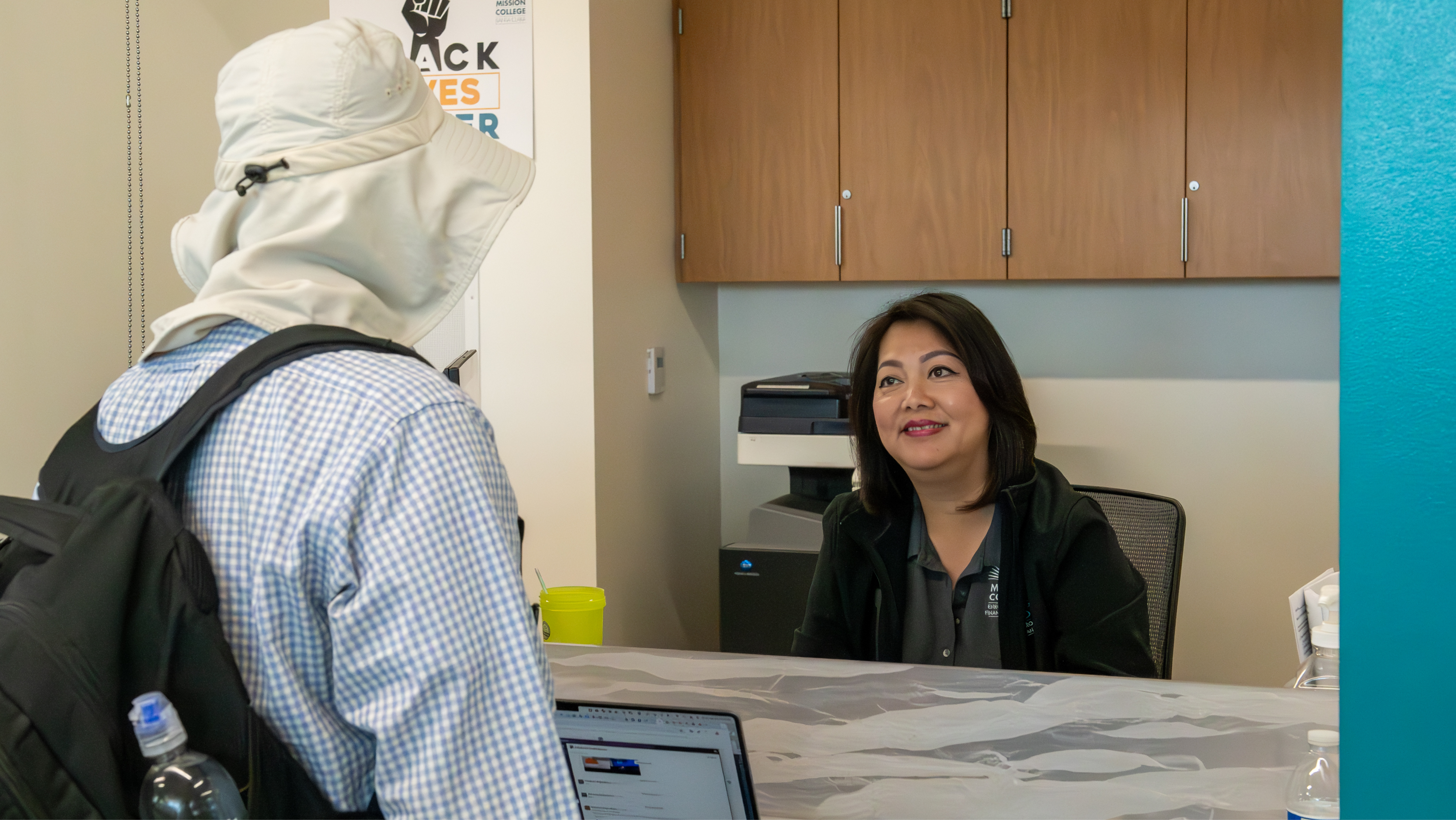 A staff member smiling warmly at a student wearing a sun hat at a campus administrative counter.