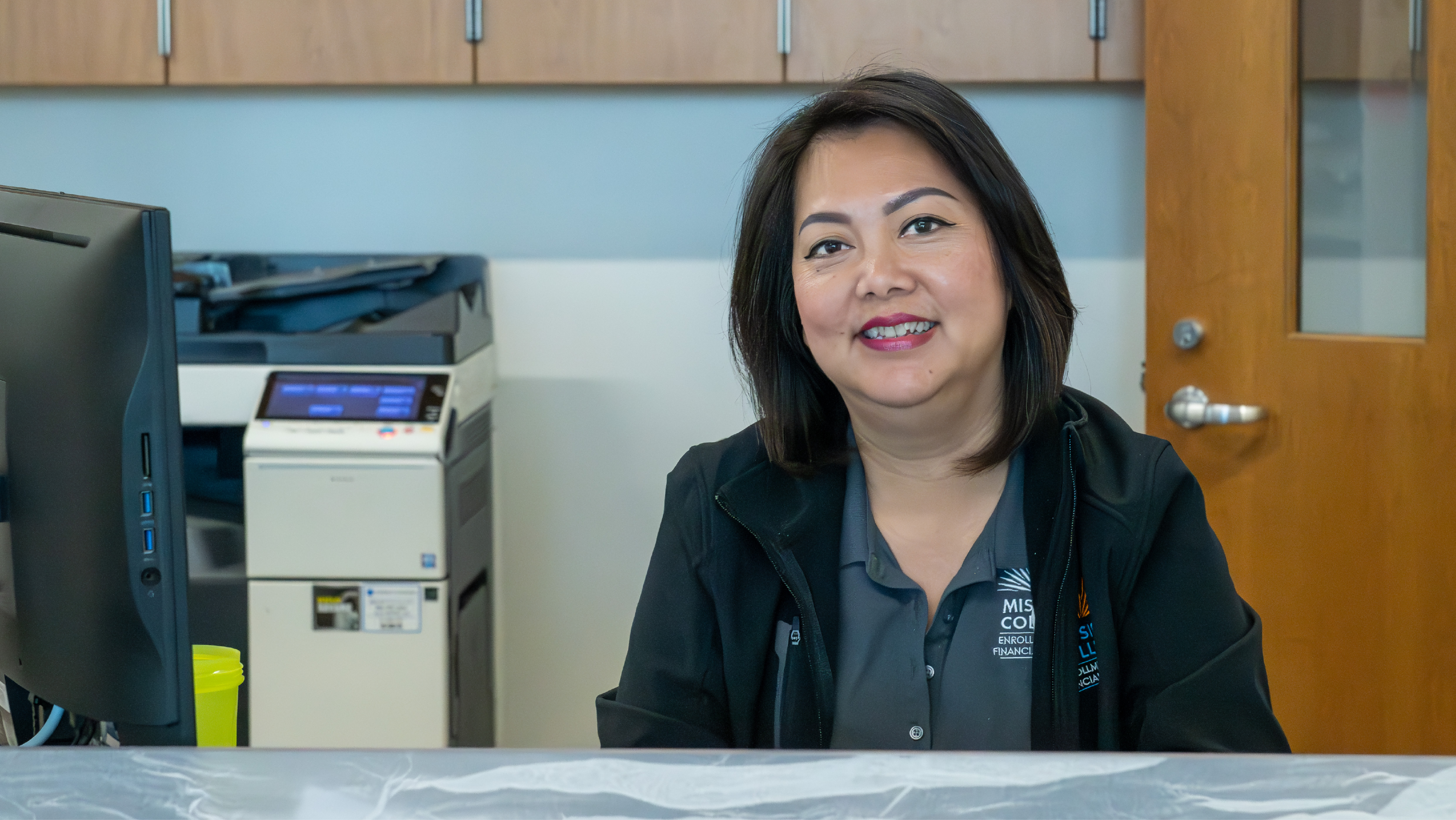 A portrait of a smiling female staff member in a gray Mission College polo seated at an administrative desk.