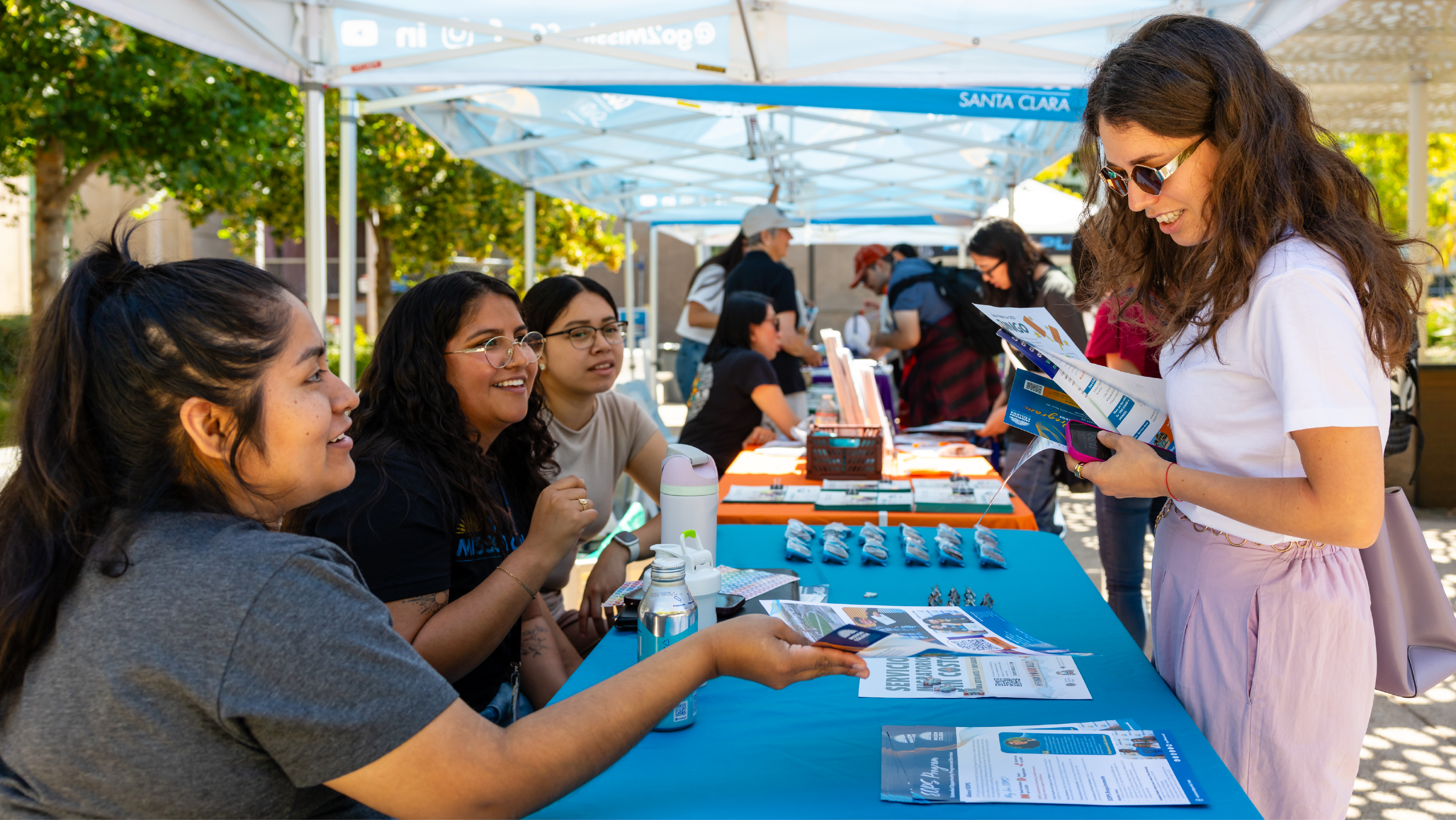 A student reviewing informational flyers at an outdoor campus canopy table hosted by smiling student representatives.