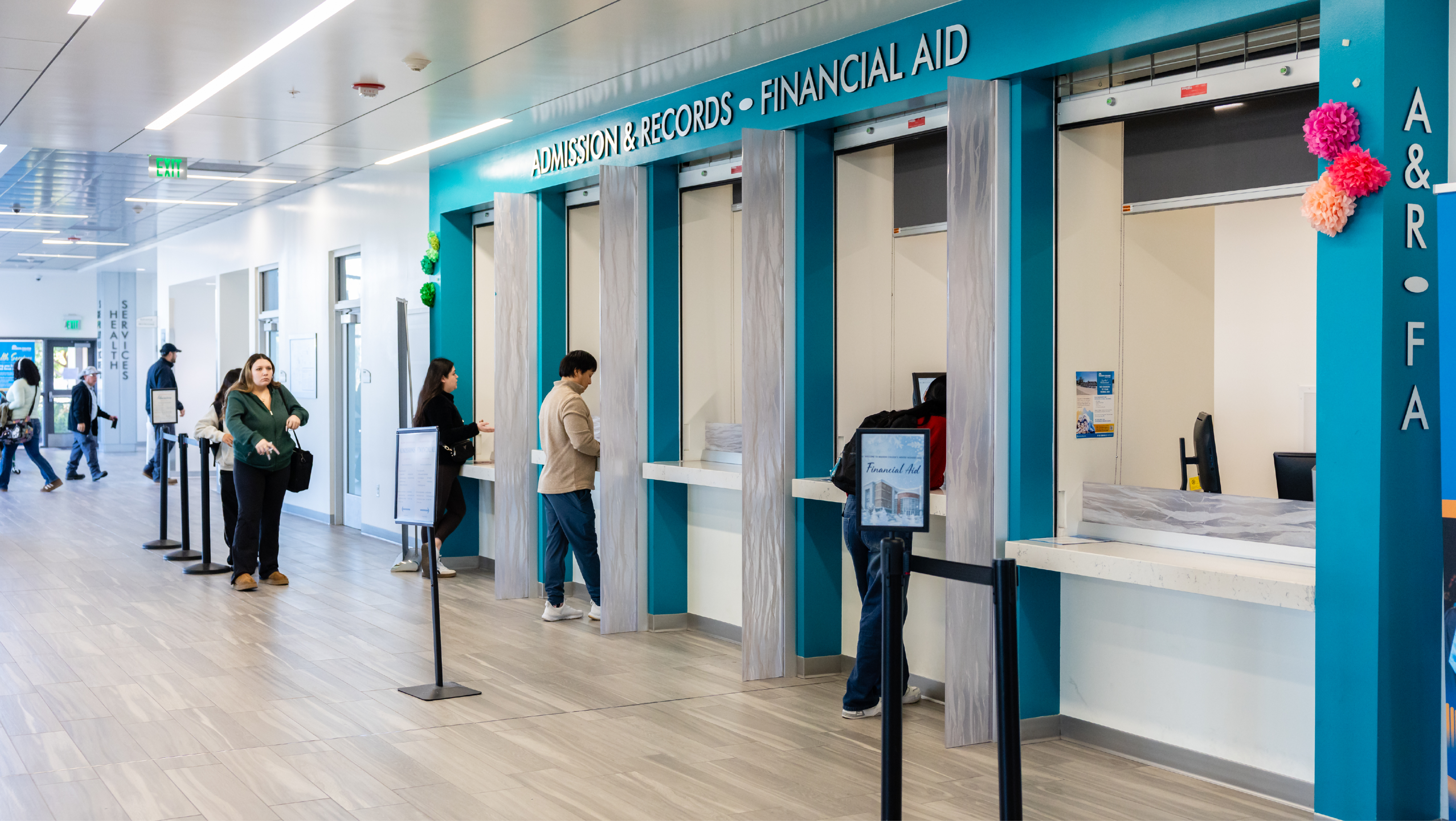 Students standing at bright blue Admissions, Records, and Financial Aid service windows in a spacious hallway.