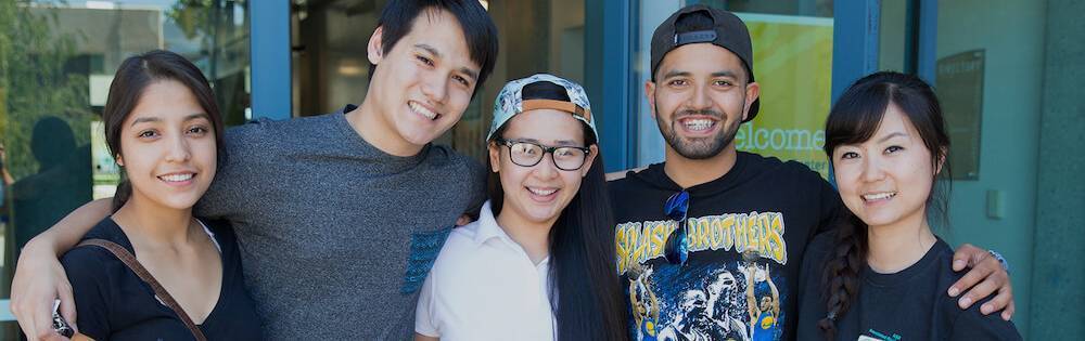 Four students with their arms around each other in front of a building on Mission campus.