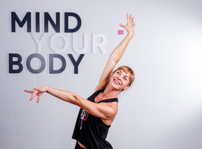 Tala Poliansky, a fitness professional with blonde hair and fair skin, wears black and performs a dance movement with outswept arms. Behind her, a sign reads "Mind Your Body."