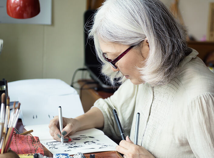 Older woman of Asian descent works on an art project at her desk. She has bobbed silver hair and wears glasses.