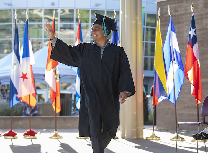 Older Latino man wears a cap and gown with a bright multi-colored sash as he walks the stage at Celebrando commencement ceremony.