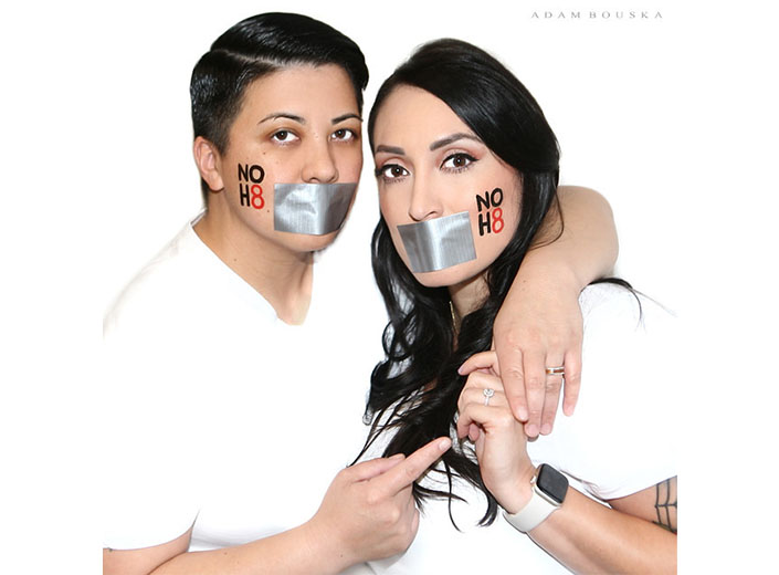 noh8 picture of a non-binary/masculine person  and a woman with long dark hair posing against a white background. They have gray masking tape over their mouths.