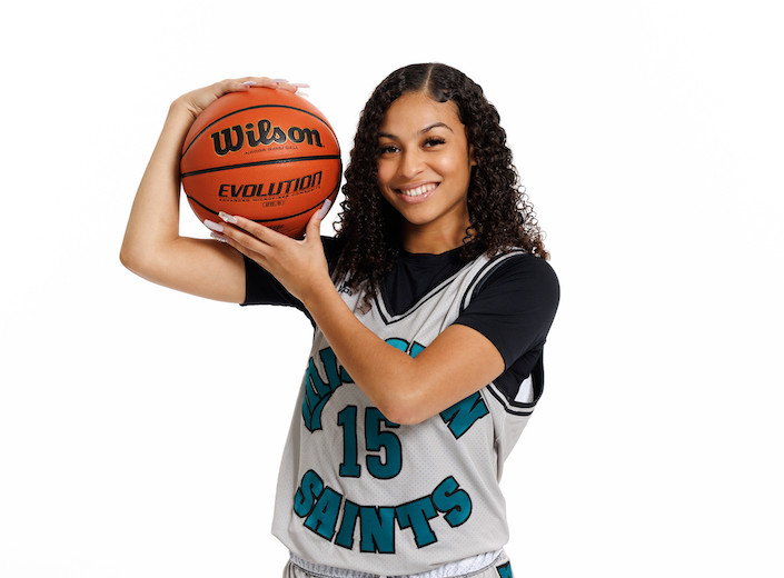 Female basketball player in MC jersey, light brown skin, and curly dark hair holds a basketball on her shoulder and smiles.
