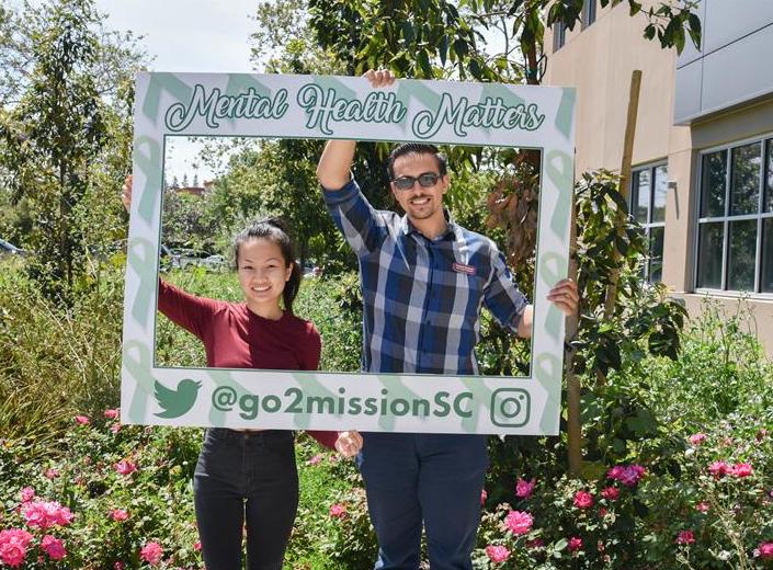 A young woman and a young man stand behind a photo frame prop that reads "mental health matter".