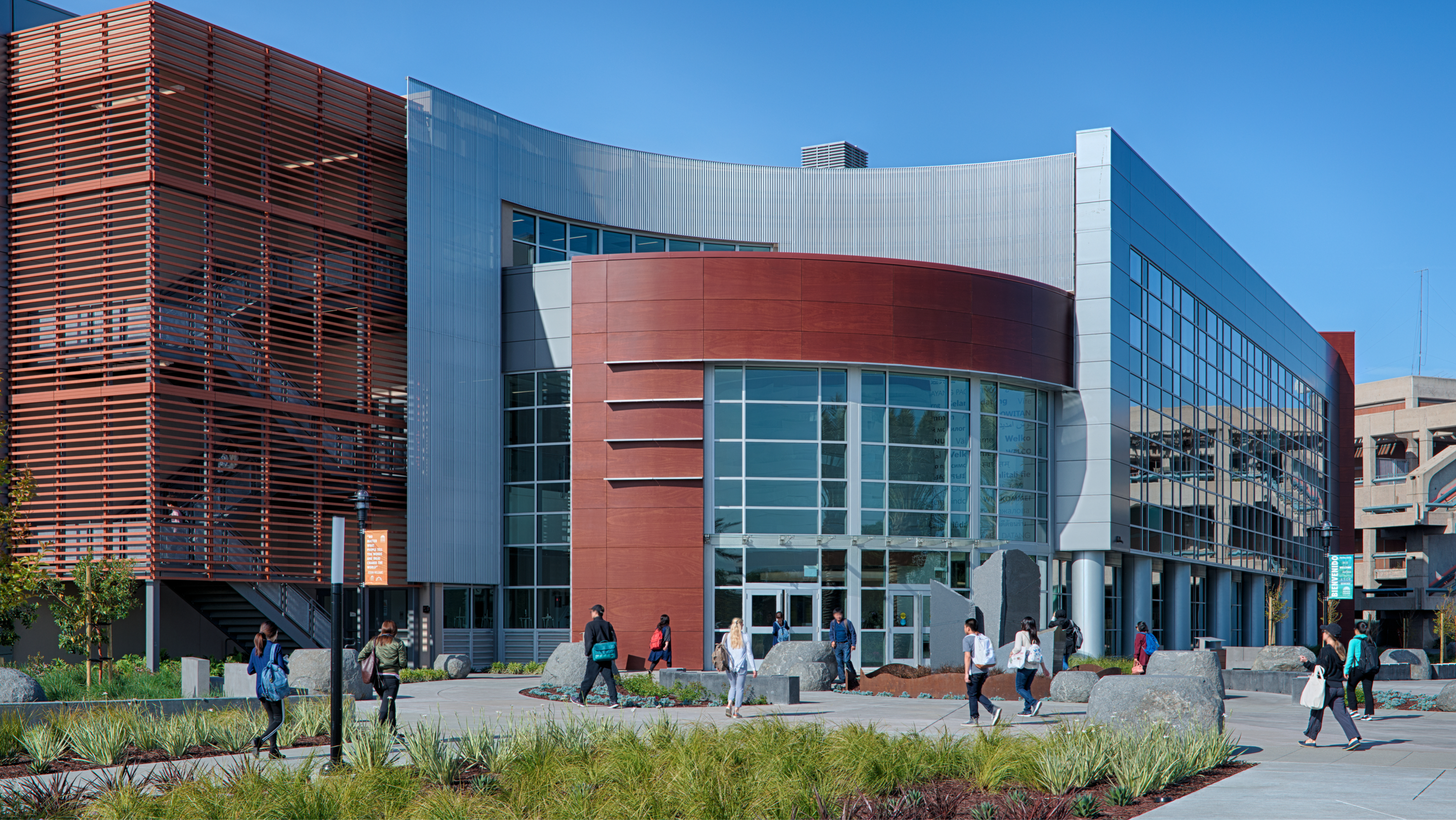 Exterior view of a modern, multi-story Mission College campus building with curved glass windows, wood-paneled accents, and students walking along the concrete plaza out front.