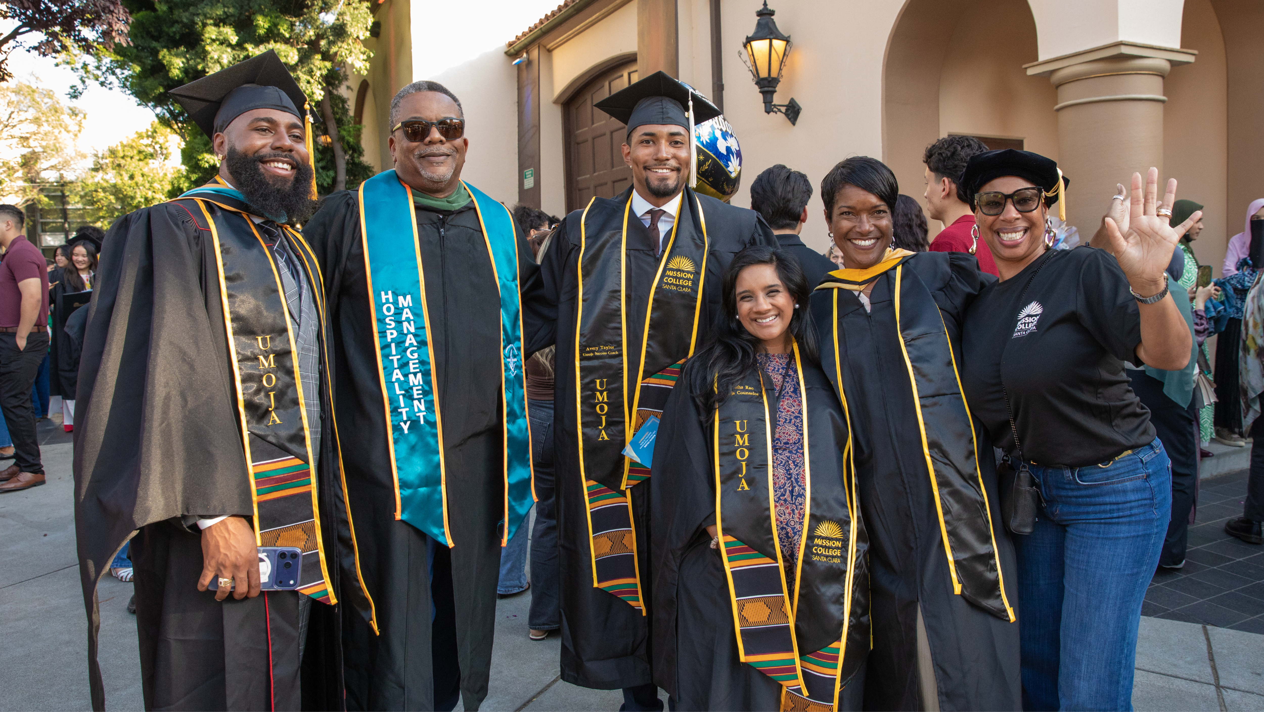 Group of Mission College graduates and faculty celebrating outside in black caps and gowns with Umoja and Hospitality Management stoles, smiling and waving at the camera.
