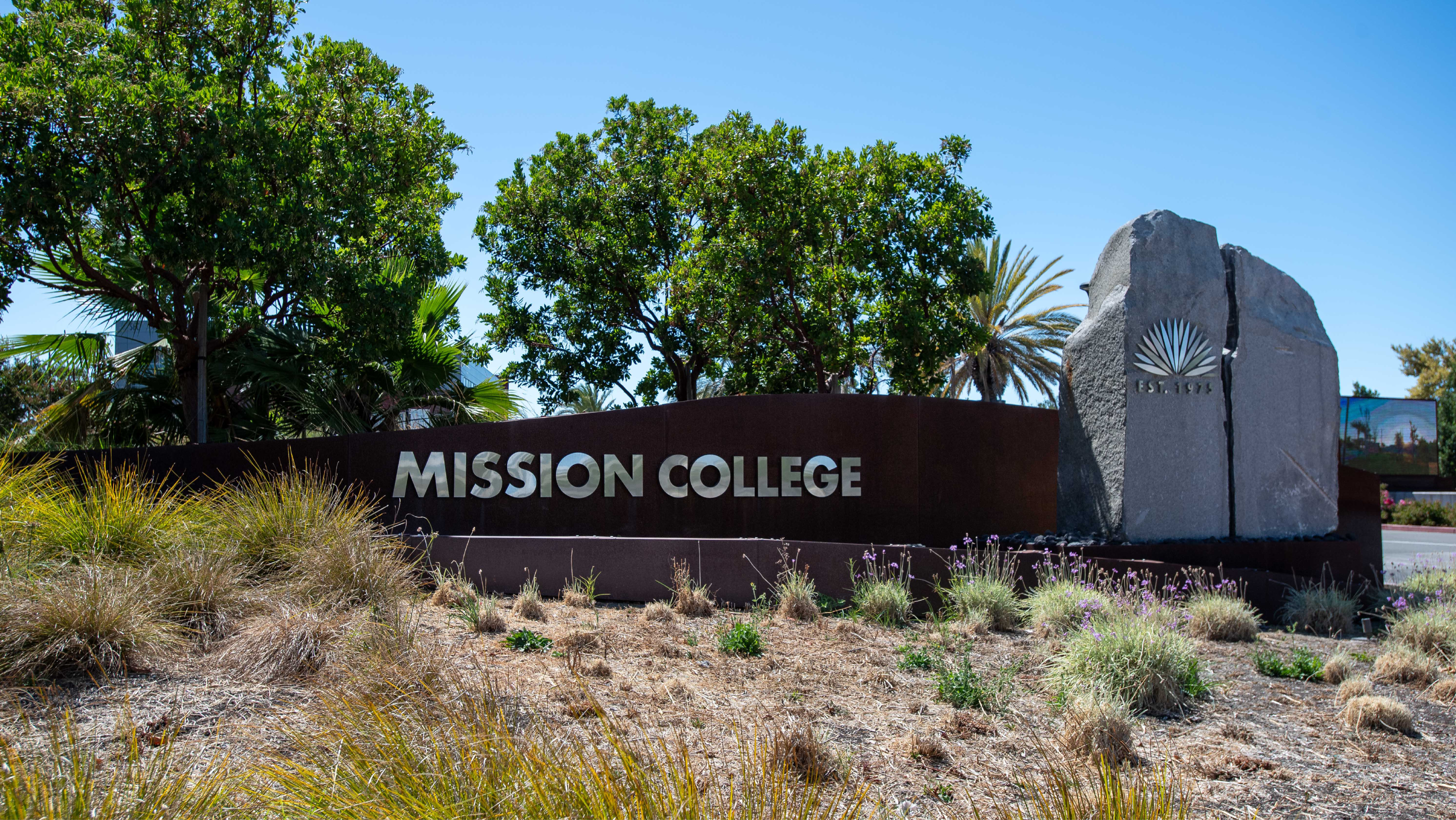Mission College entrance sign featuring metallic lettering on a dark wall next to a large stone monument carved with the college emblem and "Est. 1975."
