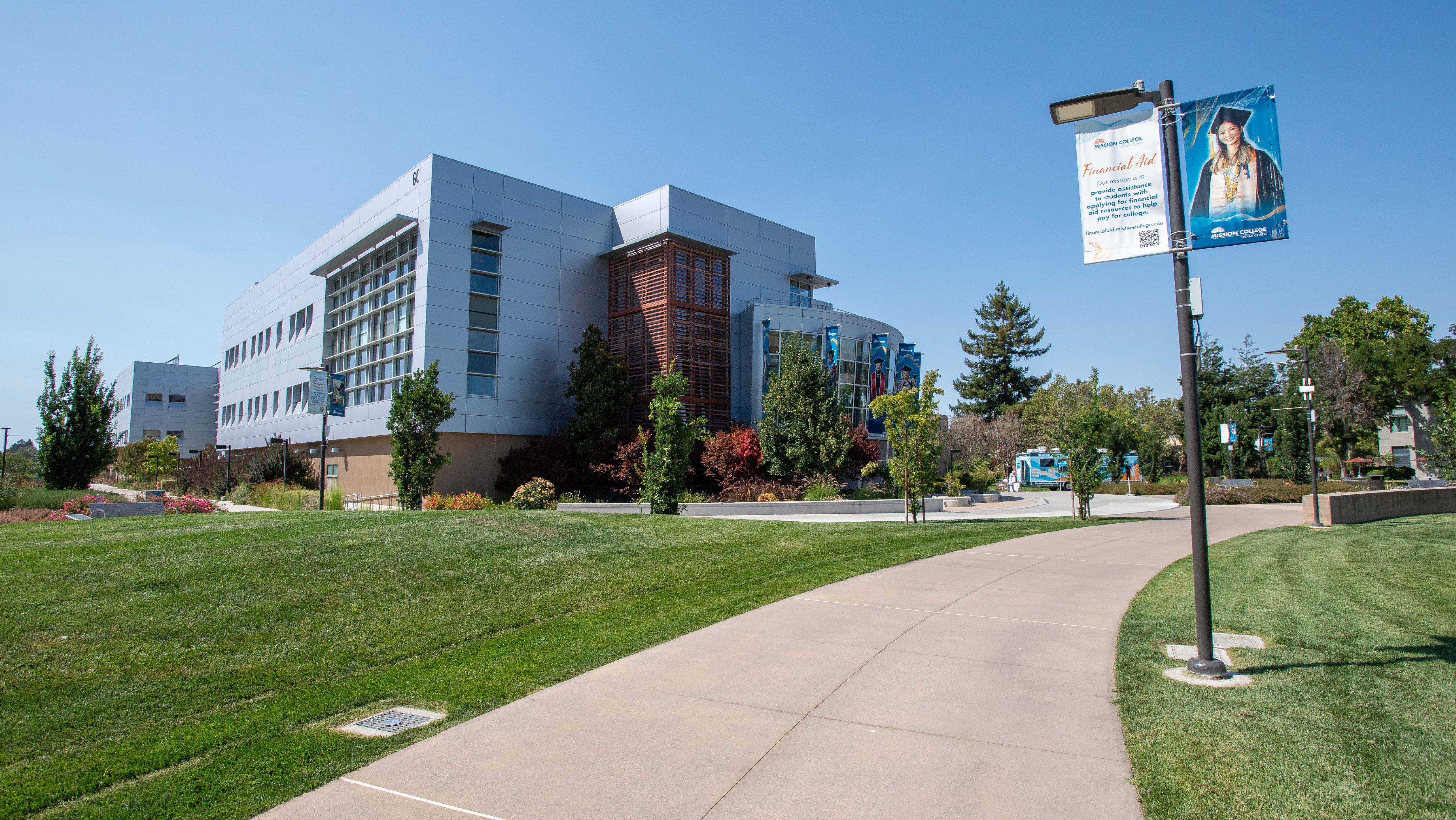 Wide view of the Mission College campus showing a curved concrete walkway, manicured green lawns, trees, modern academic buildings, and a light pole with a promotional banner.