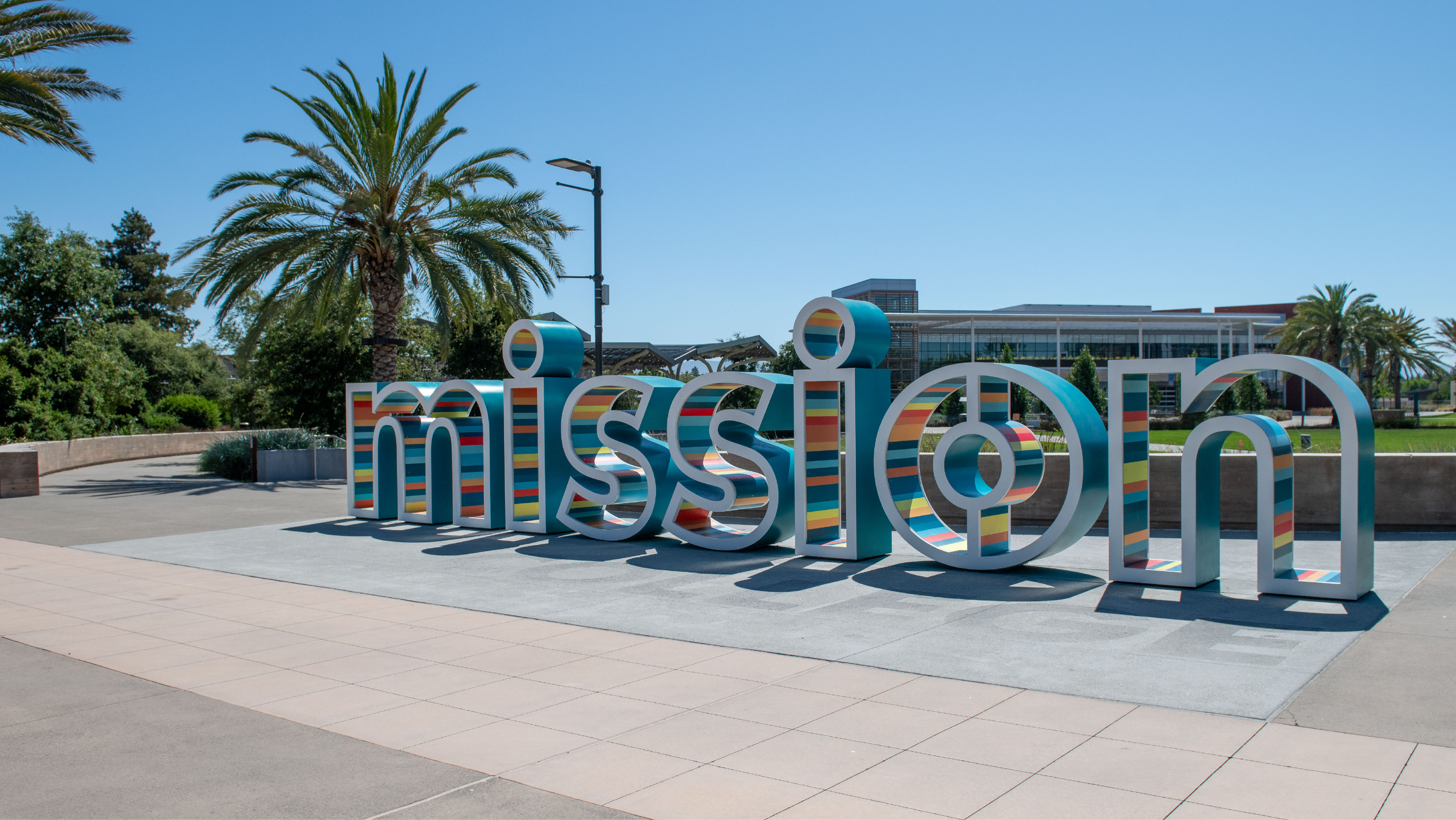 Large, colorful 3D outdoor sign spelling out "mission" in lowercase teal and striped letters on a paved campus plaza.