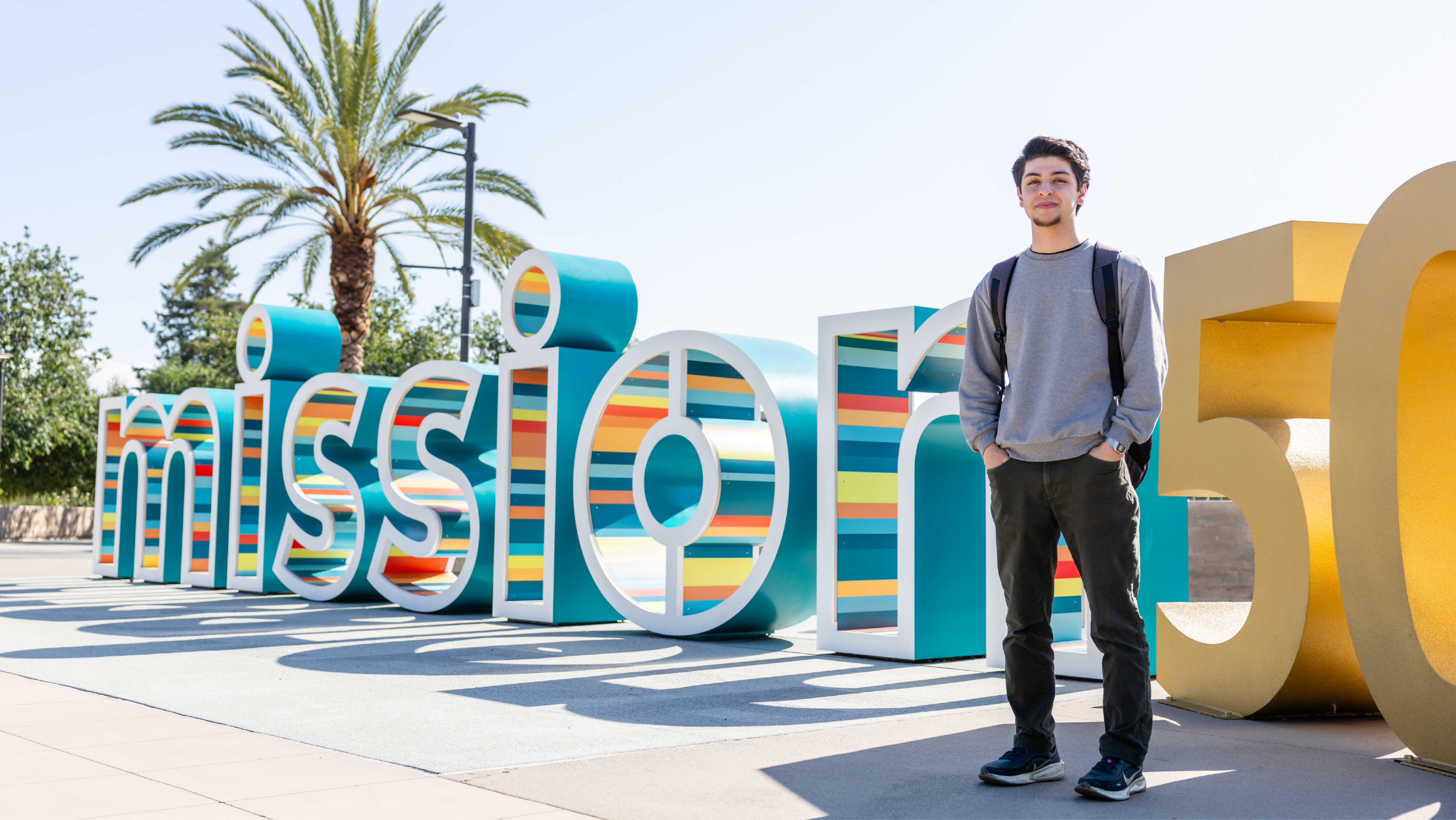 A male student wearing a grey sweater and backpack standing outdoors next to a large "Mission 50" anniversary display with colorful 3D lettering
