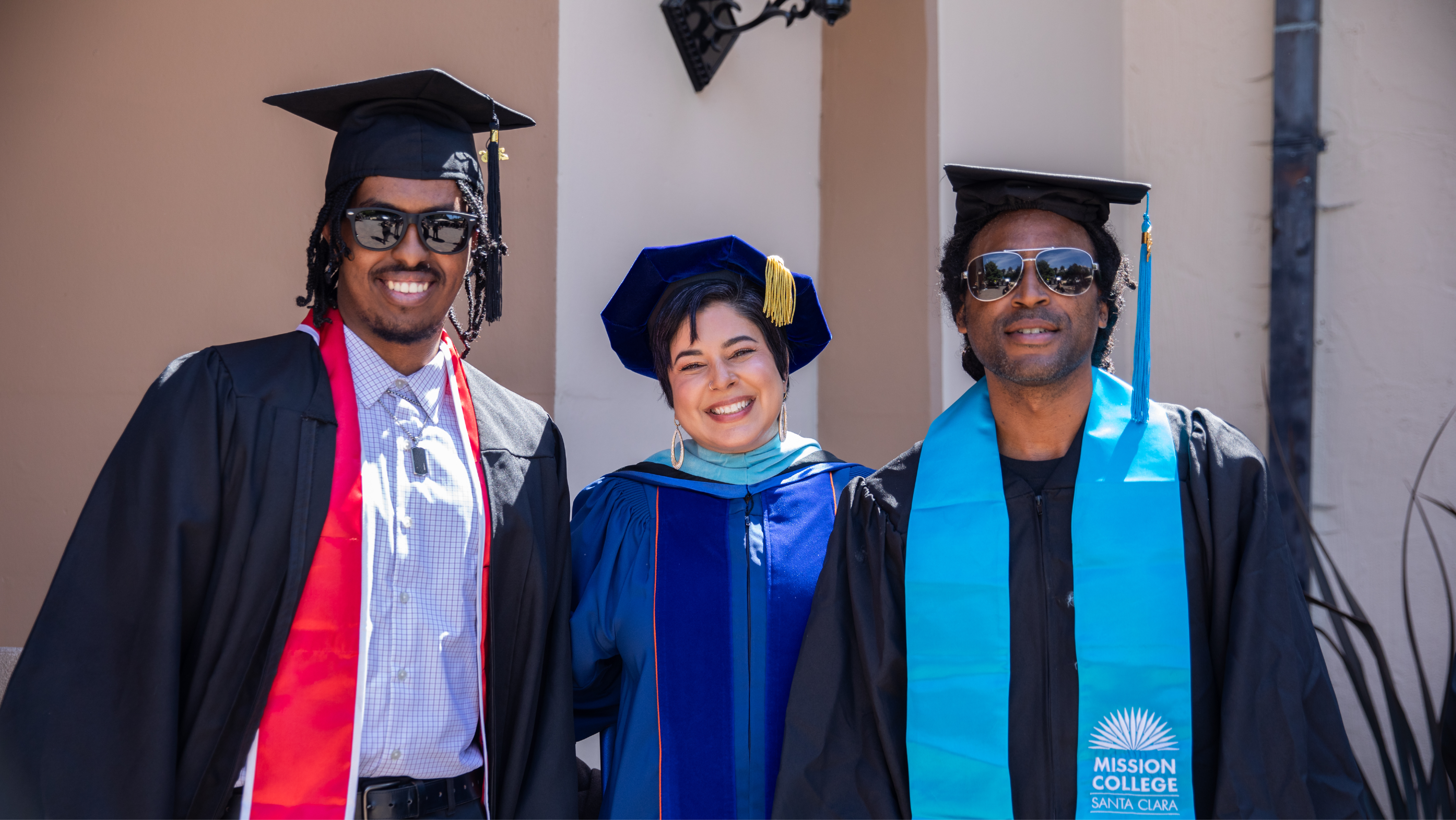 Three Mission College graduates smiling outdoors in cap and gown regalia; two male graduates wear sunglasses and teal sashes alongside a faculty member in a blue doctoral gown.