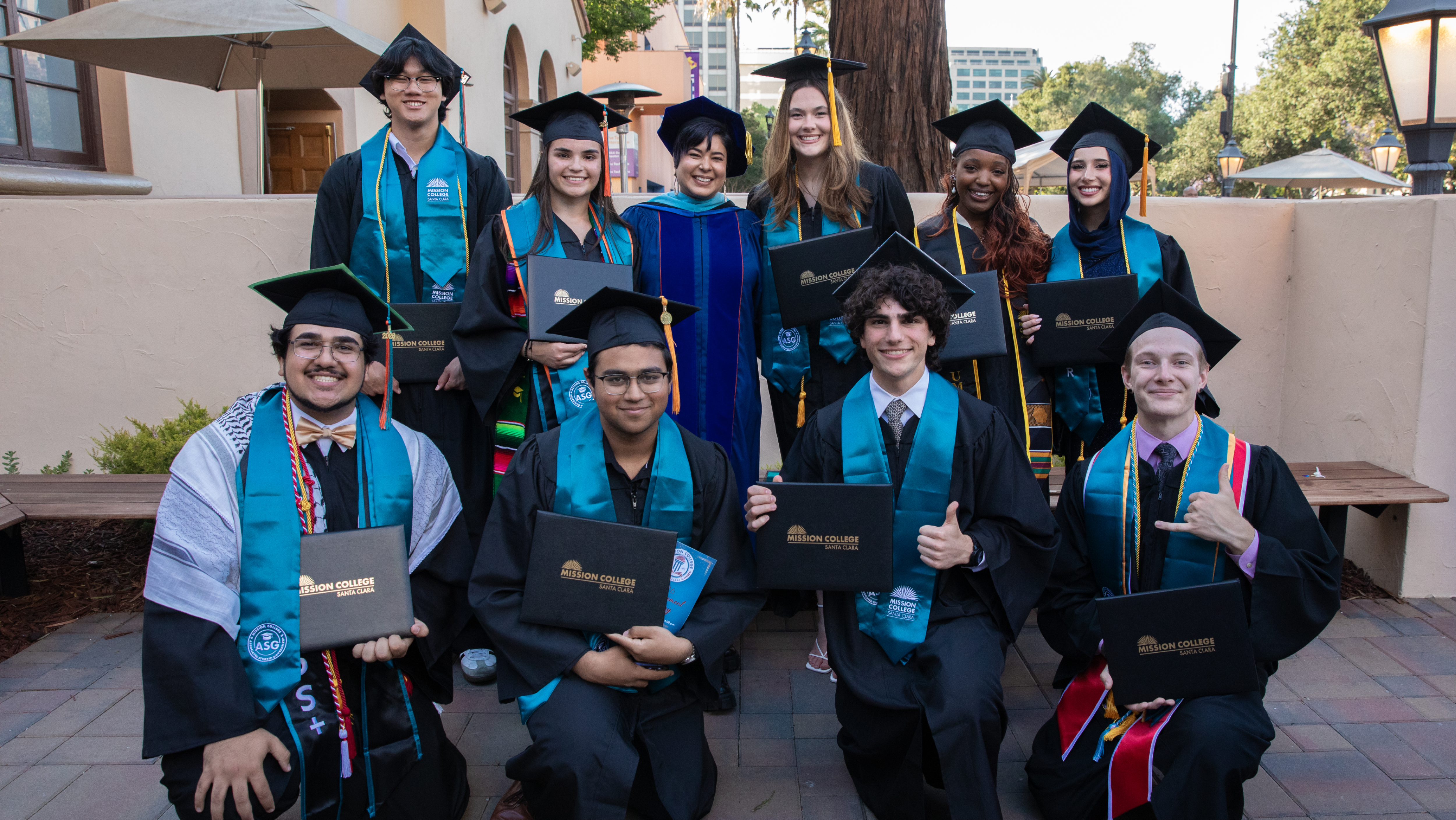 Group of Mission College graduates posing outdoors holding diploma covers, wearing black caps and gowns with teal academic stoles alongside a faculty member in a blue gown.