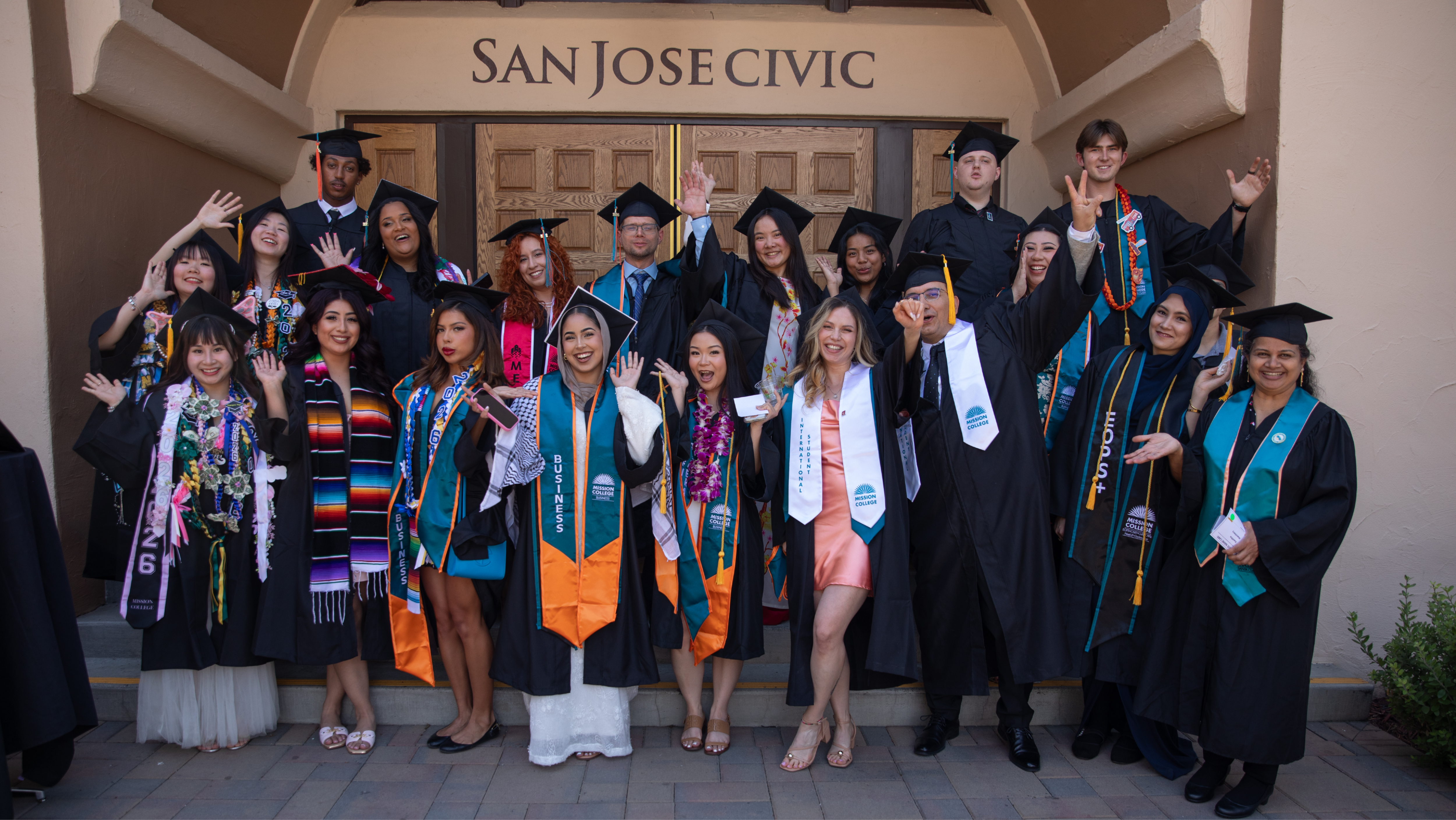 Large group of happy graduates waving and cheering in caps and gowns on the steps outside the San Jose Civic building.