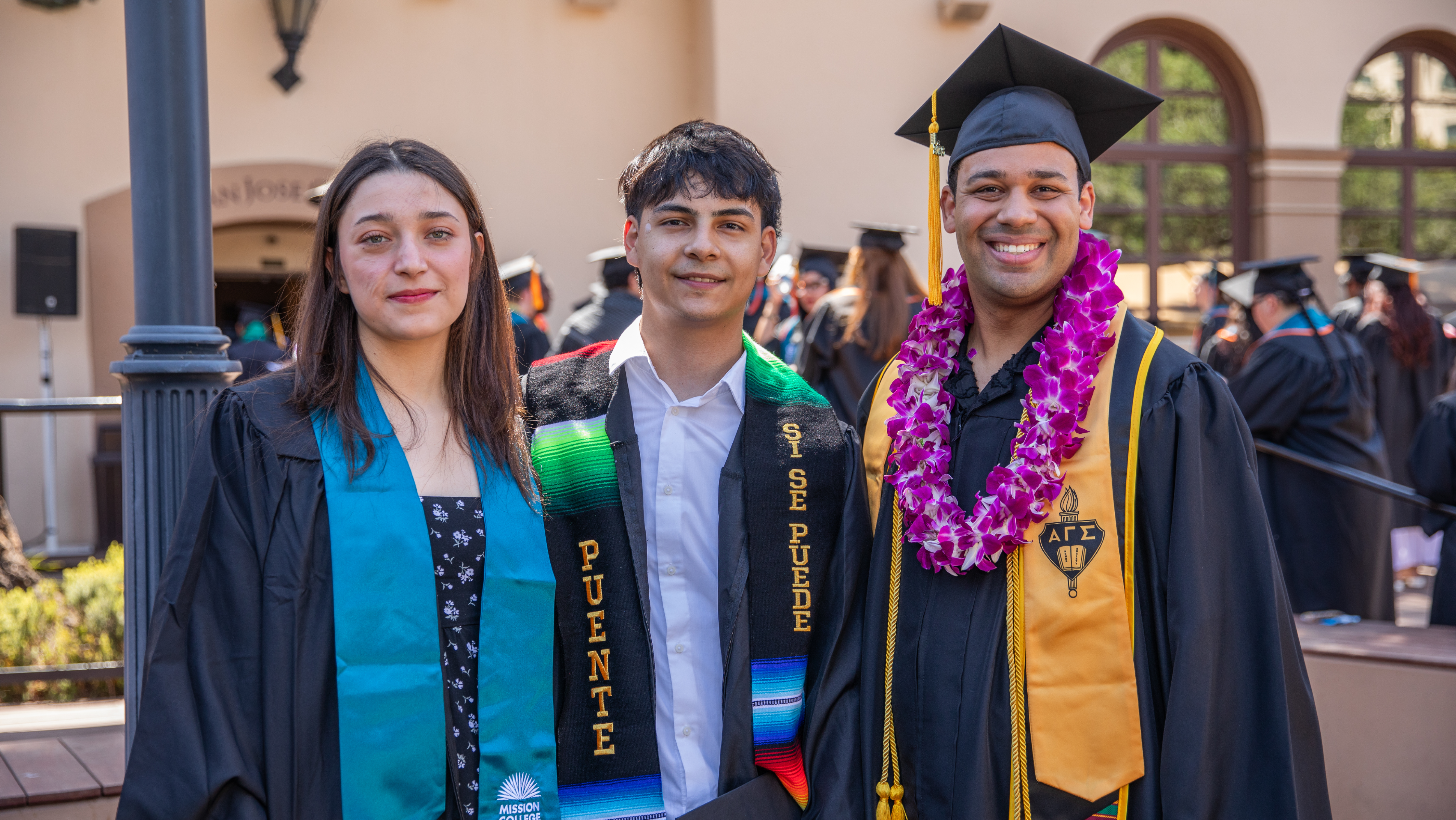 Three Mission College graduates in caps and gowns posing together outdoors, wearing teal, serape, and gold honor stoles along with a purple orchid lei.