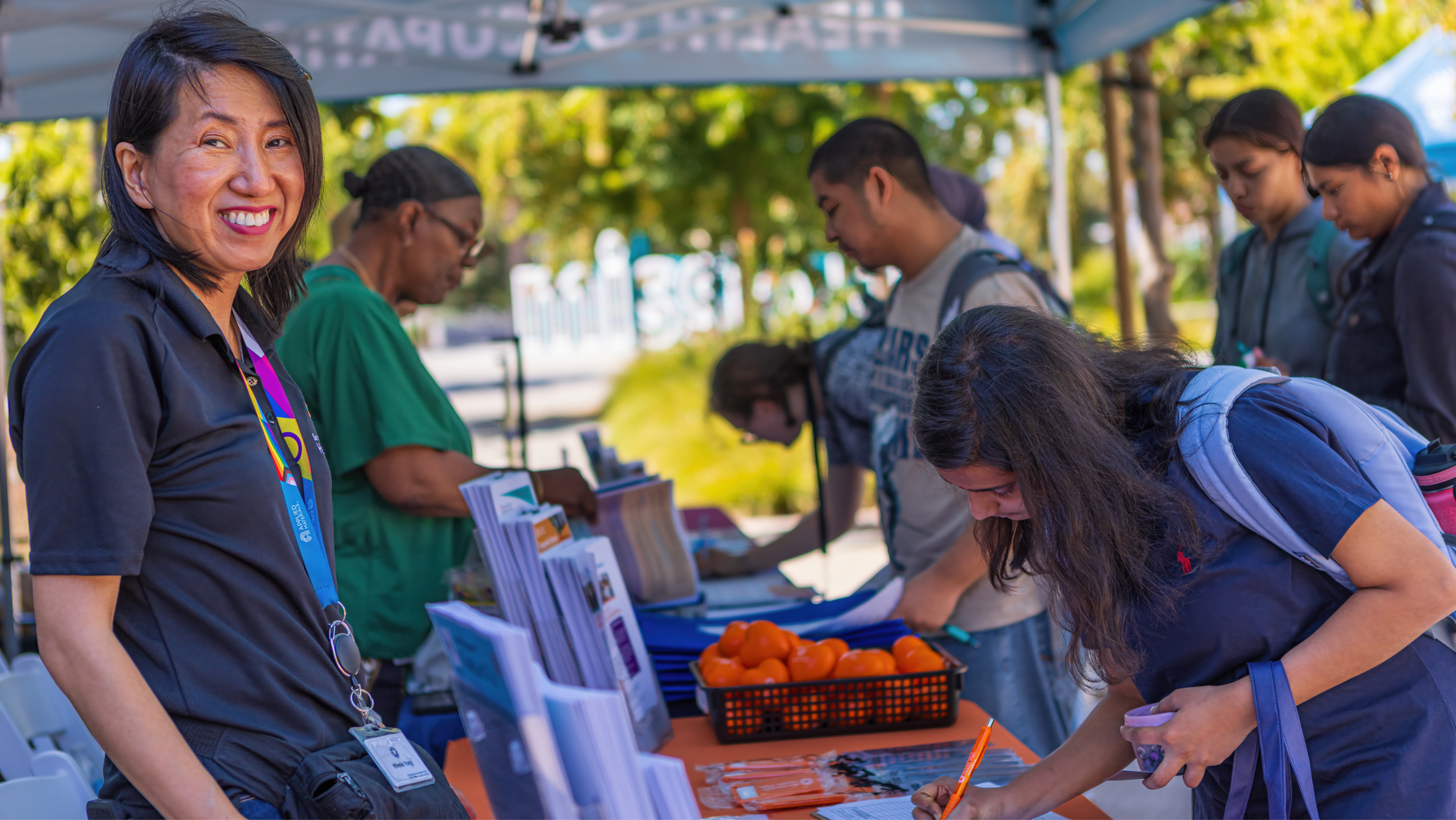 Staff member at an outdoor campus information booth smiling at the camera while a student fills out a form at a table filled with informational brochures and giveaways.